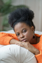 Portrait of trendy African American millennial woman in casual clothing and curly hair lying on couch, relaxing at home. Pretty thoughtful ethnic female student in orange sweater looking at camera.