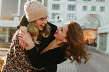 Debonair girls enjoying autumn weekend. Pretty young woman in knitted hat embracing her friend.