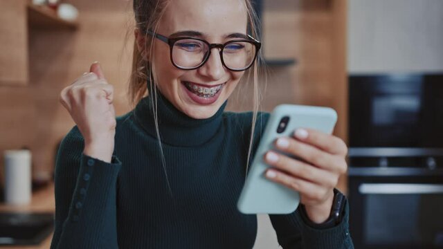 Funny young woman with glasses use phone smiling sitting near table. Message texting workplace smartphone browsing internet device. Slow motion