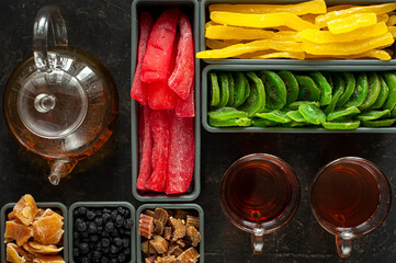 A set of different dried fruits and candied fruit in rectangular and square containers, two cups and a teapot with tea on a black background. Knolling. A healthy snack. Winter sweets. Vegan food.