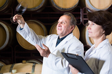 smiling worker of winery holds glass of red wine in hand and shows him to expert