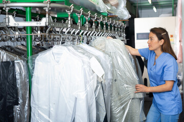 Positive female dry-cleaning worker preparing garments for collection, hanging clean clothes packed...