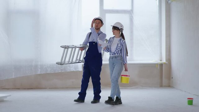 portrait of joyful man and woman in protective helmets with stepladder and bucket of paint in their hands discussing color of walls, during renovation of walls