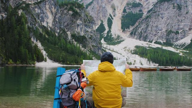 Young tourist hiker man reading map sitting by alpine lake Lago Di Braies (Pragser wildsee) in Trentino, Dolomites mountains, Italy.