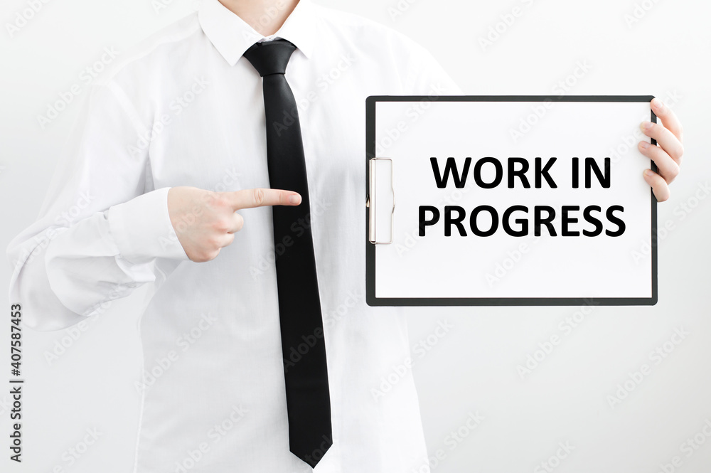 Poster man belay in a shirt holds a tablet with text work in progress written in it on a gray background. b
