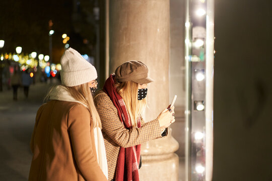 Two young women in a city at night, in winter clothes, looking at a store window. One of them takes a picture of something with her phone.