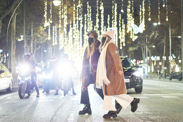 Two young women walking at a crosswalk. City crosswalk at night. In the background are several vehicles stopped at the traffic light.