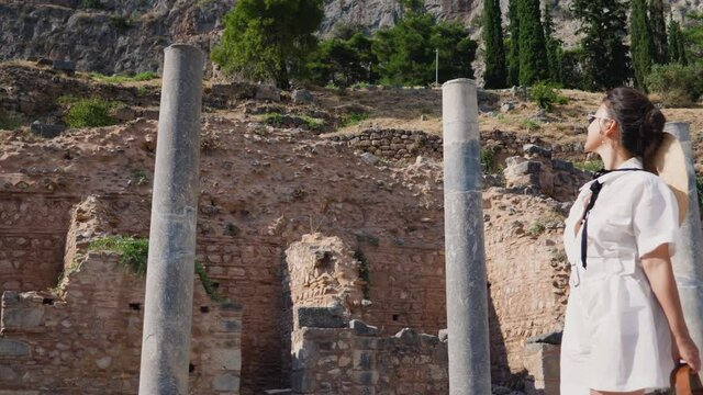 Young woman visiting ruins of temple of Apollo in Delphi, archaeological site by mount Parnassus, Greece, Europe. Large hat, fashion white dress.