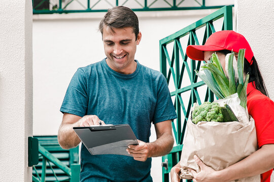 Happy man signing for receiving order from grocery store, holding clipboard and smiling. Postwoman in red uniform holding paper bag with vegetables. Food delivery service and post concept