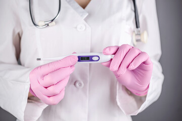 A female doctor in a white medical gown and pink gloves is holding a thermometer. Concept of health care. Closeup and gray background
