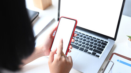 Close up view of young female holding smartphone getting message with confirmation making transaction on laptop computer.