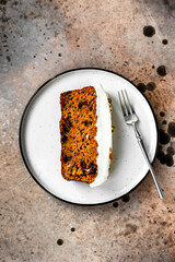 A piece of a carrot cake with cranberries, walnuts and cream cheese frosting, a dessert fork, on a white plate. Close-up top view
