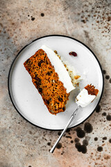 A piece of a carrot cake with cranberries, walnuts and cream cheese frosting, a dessert fork, on a white plate. Close-up top view