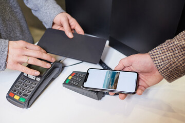 A close-up hand of a male customer who is holding a smartphone while paying through a digital wallet using NFC. A cashier accepts payment transaction at a contactless-capable point-of-sale terminal
