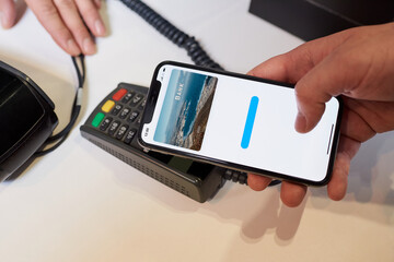 A close-up hand of a male customer who is holding phone while paying through a digital wallet using NFC in a store. Cashier accepts payment transaction at a contactless-capable point-of-sale terminal