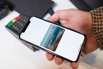 A close-up hand of a male customer who is holding a smartphone while paying through the digital wallet using NFC technology for his purchases in a store.