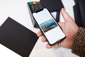 A close-up hand of a male customer who is holding a cellphone while paying through a digital wallet using NFC for his purchases in a store. Transaction at a contactless-capable point-of-sale terminal