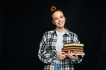 Cheerful young woman college student holding book and looking at camera on isolated black background. Pretty redhead lady model wearing casual fashion clothes emotionally showing facial expressions.