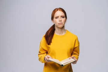 Obraz premium Confused displeased young woman college student holding book and crying on gray isolated background. Pretty redhead lady model wearing yellow sweater emotionally showing facial expressions.
