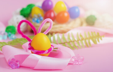 A yellow Easter egg with pink bunny ears and a satin ribbon in the foreground against a background of colorful eggs in a wicker nest against a pink background. Horizontal format, banner. Copyspace
