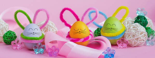A set of colorful eggs with a drawn rabbit face and ears lie in a satin ribbon with ornaments on a pink background. Horizontal banner. Postcard for the Easter holiday. Copyspace