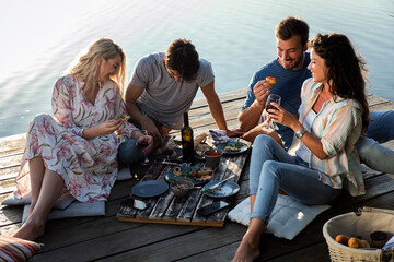 Group of friends having fun on picnic near a lake, sitting on pier eating and drinking wine.