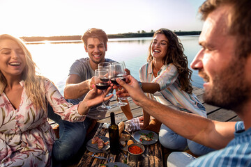 Group of friends having fun on picnic near a lake, sitting on pier eating and drinking wine.