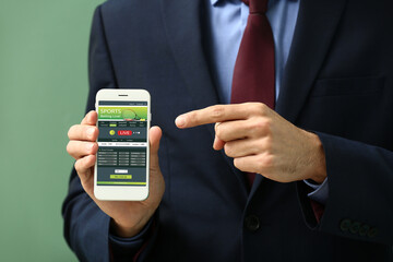 Young man with open page of online betting site on screen of mobile phone against color background,...