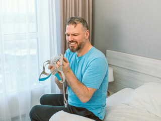 Unhappy shocked man with chronic breathing issues surprised by using  CPAP machine sitting on the bed in bedroom. Healthcare, CPAP, Obstructive sleep apnea therapy, snoring concept