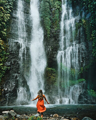 Young woman standing in front of a very big and beautiful waterfall. Dancing next to sekumpul waterfall in bali, indonesia.  - obrazy, fototapety, plakaty