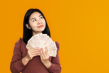 Asian woman hand holding Thai banknotes with happy face smiling over yellow background dreaming of spending money