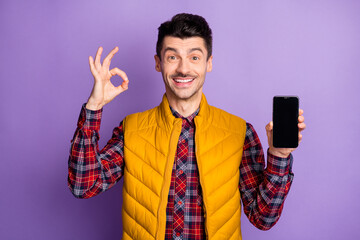 Photo of cute funny young man wear yellow sleeveless jacket holding modern gadget showing okey...