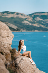 Joyful woman travel mountains. Authentic blond female relax in picturesque landscape. A girl with developing natural hair is wearing a white top and denim shorts.