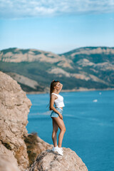 Joyful woman travel mountains. Authentic blond female relax in picturesque landscape. A girl with developing natural hair is wearing a white top and denim shorts.