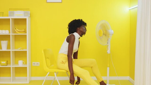 Young Black Woman Is Sitting In Front Of Fan, Trying To Chill Out In Hot Yellow Room, Feeling Bored And Chewing Bubble Gum, Looks Into A Camera, Creative Video Concept, Slow Motion.