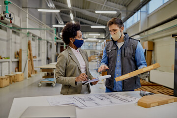 Black businesswoman and woodworker with face masks talking analyzing processed wood in a factory