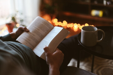 A book in man's hand, a man sitting on a couch with a cup of tea, warm candid light in a cosy...