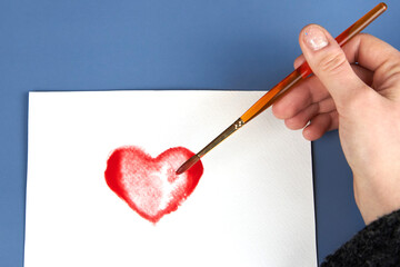 A woman's hand draws a red heart shape in watercolor on white paper, Valentine's Day theme.