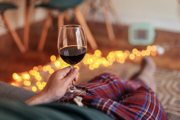 Man in check pyjamas holding a glass of red wine, cosy living room with candid light