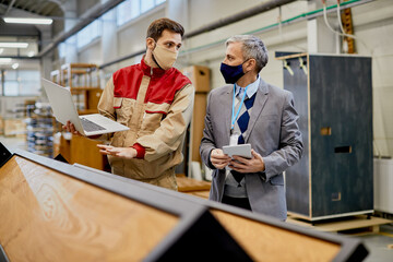 Male worker using laptop while communicating with company manager at wood factory during coronavirus pandemic.