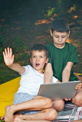 Children play in nature with a laptop. The boys are resting in a tent camp. Children sit on the carpet on the grass in summer.