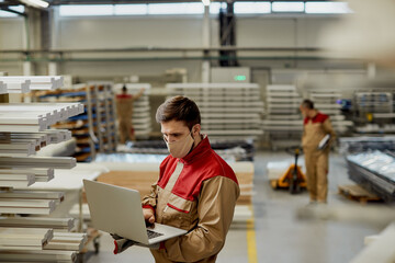 Male worker with face mask using computer while working at carpentry workshop.