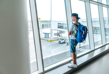 Lonely teen solo traveler with backpack standing in the empty airport passenger transfer hall in protective face mask and looking out large windows. Traveling in worldwide pandemic time concept image