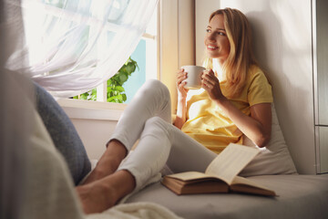 Beautiful young woman with cup of drink near window at home