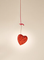 Red glittery heart hooked with piece of metal hanging on pink thread on a beige background Shoot...
