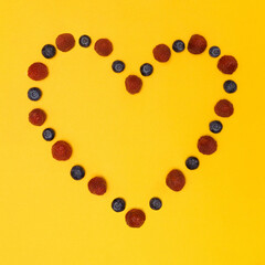 Funky fruit bordered heart shaped copy space made of fresh berry fruit maroon colored raspberries and dark blue blueberries. Flat lay pastel yellow orange background.