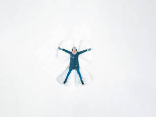 Young beautiful woman in a gray jacket, hat and gloves, blue jeans, boots makes a snow angel in the snow, a view from a drone. Snow fun, entertainment