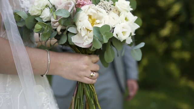Young married couple of caucasian newlyweds stand among nature and the bride with wedding ring and bracelet on her hand holds delicate luxurious elegant bouquet with eustoma pastel colors and greens