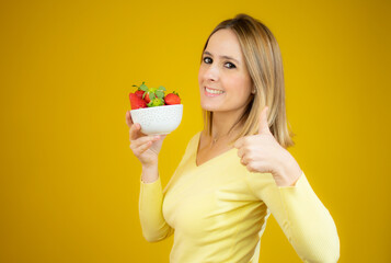 Beautiful cheerful young woman standing and holding glass bowl with strawberries with thumb up over yellow background