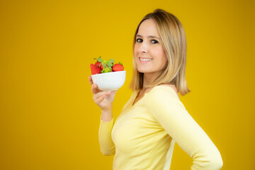 Beautiful cheerful young woman standing and holding glass bowl with strawberries over yellow background